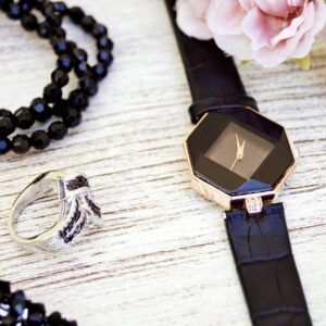 Close-up of luxurious black jewelry and watch with pink flowers on a wooden surface.