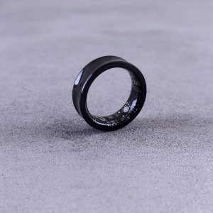 A modern black smart ring with visible circuitry resting on a textured concrete surface.